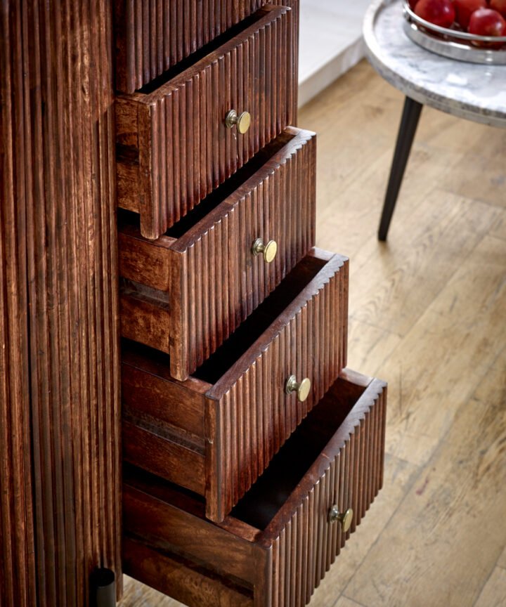 Close-up of a rich brown Opal Mango Wood chest of drawers with vertical grooves and small brass knobs, showcases stacked open drawers in a rustic interior scene.
